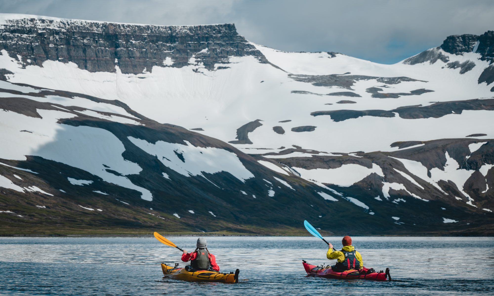Zwei Seekajaks paddeln in einem Fjord auf einen schneebedeckten Berg zu.